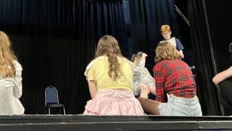 Young Company are onstage, watching two members rehearsing a script.